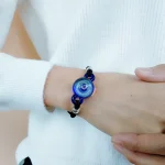 Pulseira com elemento de vidro azul e detalhes em preto e prata, usada no pulso de uma pessoa com roupa branca de manga comprida.