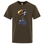 T-shirt marrom com um padrão de astronauta flutuando segurando um balão formado por várias planetas coloridas, criando uma cena espacial lúdica e imaginativa.