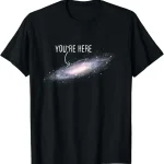 T-shirt galáxia com texto "You're Here" em branco sobre fundo preto, representando uma galáxia espiral com uma mensagem humorística e cósmica.