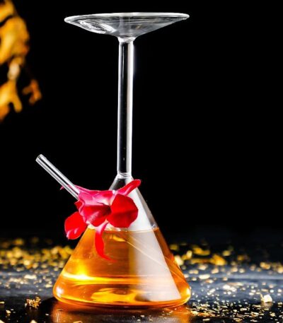 Béchier de laboratoire avec verre à cocktail suspendu, contenant un liquide orange, décoré d'une fleur rouge, sur une surface noire avec des éclats dorés.