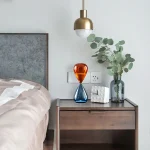 Vaso em vidro laranja e azul sobre uma mesa de cabeceira moderna com planta verde e lâmpada suspensa dourada.