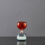 Balança de ampulheta em vidro vermelho e transparente com areia branca.
