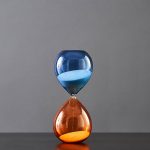 Pingente em forma de ampulheta com areia azul e laranja, simbolizando o tempo e a ciência, perfeito para decoração ou como acessório científico original.