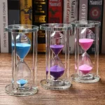 Ampulheta colorida em vidro com três tubos contendo areia azul, roxa e rosa, colocada sobre uma superfície de madeira com livros ao fundo.
