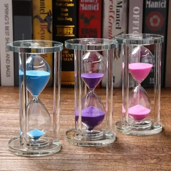 Ampulheta colorida em vidro com três tubos contendo areia azul, roxa e rosa, colocada sobre uma superfície de madeira com livros ao fundo.