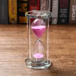 Ampulheta de vidro com areia rosa vibrante, símbolo do tempo que passa, colocada sobre uma superfície de madeira com uma estante ao fundo.
