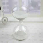 Ampulheta em vidro com areia branca, símbolo do tempo que passa, colocada sobre uma superfície clara com um fundo minimalista.