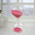 Ampulheta em vidro transparente com areia rosa, símbolo do tempo que passa, colocada sobre uma superfície clara com um fundo minimalista.