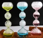 Balanço de vidro colorido com bolhas de ar, composto por três segmentos em forma de bola, cada um preenchido com líquido colorido (amarelo, azul, rosa) e bolhas de ar, sobre fundo preto.