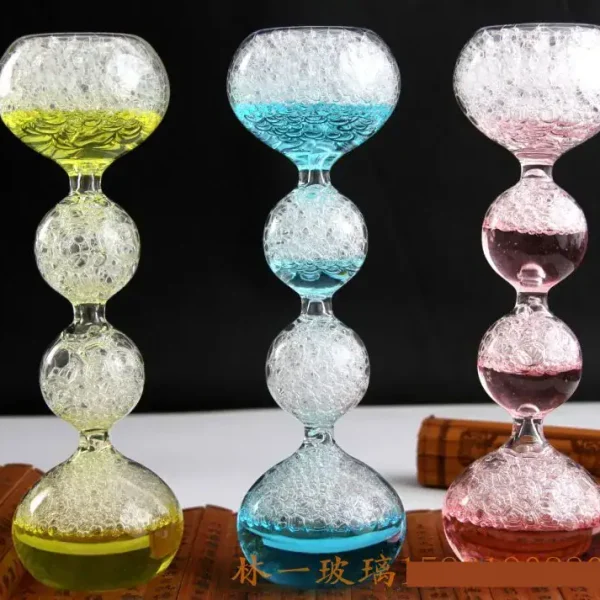 Balanço de vidro colorido com bolhas de ar, composto por três segmentos em forma de bola, cada um preenchido com líquido colorido (amarelo, azul, rosa) e bolhas de ar, sobre fundo preto.