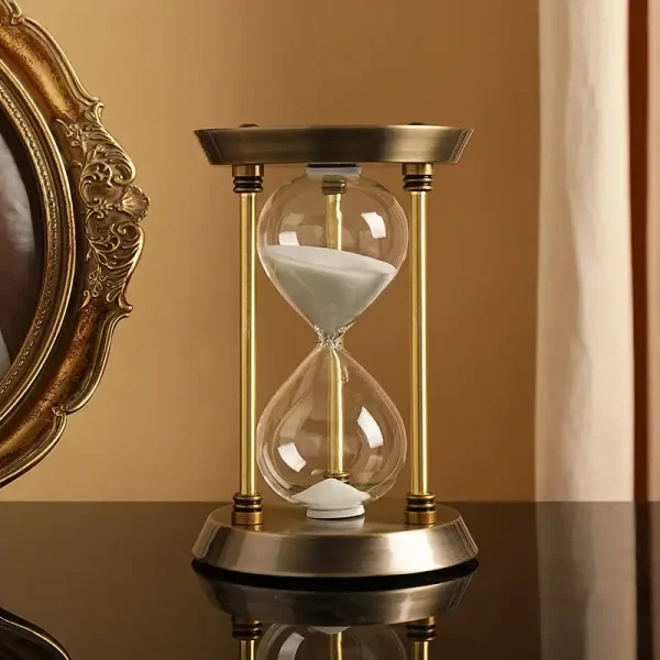 Ampulheta decorativa em vidro com areia branca, enquadrada por uma estrutura em metal dourado, colocada sobre uma superfície de madeira escura, em um interior elegante.