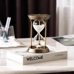 Ampulheta de decoração em latão e vidro, colocada sobre um livro intitulado "WELCOME" em um ambiente de escritório ou sala de estar.