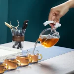 Pessoa servindo chá quente em xícaras de vidro, com um bule de vidro ao lado, sobre uma mesa de madeira.
