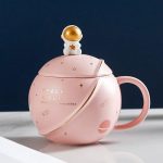 Vaso decorativo em forma de caneca de chá com motivo espacial, incluindo uma tampa com um pequeno astronauta dourado. Perfeito para uma decoração original em uma mesa ou prateleira.