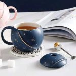 Caneca de cerâmica azul com motivos de astros, acompanhada de um mouse de computador combinando, sobre uma mesa branca com um livro aberto ao fundo.