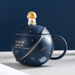 Caneca decorativa em forma de nave espacial com um pequeno astronauta no topo, decorada com motivos planetários e estrelas, ideal para amantes de astronomia e decoração científica.