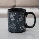 Caneca de cerâmica preta com ilustrações das constelações do zodíaco: Gêmeos, Sagitário, Leão, Áries, e mais, com nomes em francês.