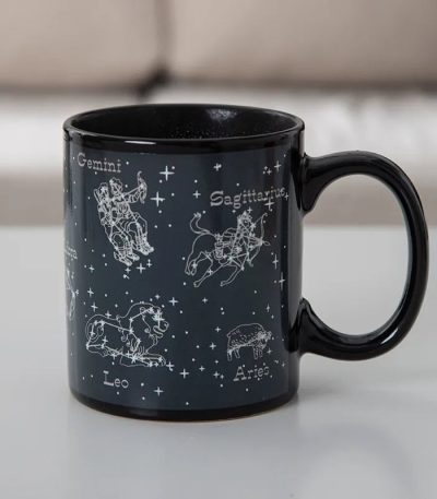 Caneca de cerâmica preta com ilustrações das constelações do zodíaco: Gêmeos, Sagitário, Leão, Áries, e mais, com nomes em francês.