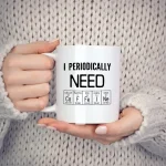 Caneca branca com mensagem "I Periodically NEON" e símbolos de elementos químicos, segurada por uma pessoa com suéter bege, sobre fundo neutro.
