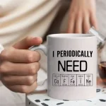 Caneca branca com texto "I PERIODICALLY NEED" e tabela periódica ilustrando elementos químicos, perfeita para os amantes de ciência e química.