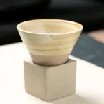 Vaso em cerâmica bege com acabamento liso, colocado sobre um pedestal de concreto branco, ideal para decoração interior ou como peça de coleção científica.