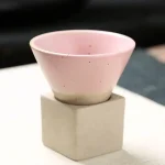 Taça em cerâmica rosa pastel colocada sobre um pedestal de concreto, ideal para decoração de estilo científico ou moderno.
