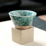 Pequeno pote em cerâmica verde com efeito marmoreado, colocado sobre um pedestal branco, ideal para decoração científica ou botânica.
