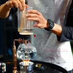 Pipeta de vidro manipulada por uma mão durante uma experiência científica, com um balão de laboratório contendo um líquido transparente, ilustrando a precisão e técnica em química.