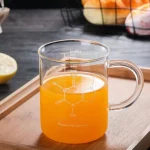 Copo de química em vidro transparente cheio de um líquido laranja, colocado sobre uma tábua de madeira, com frutas ao fundo.