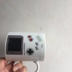 Console de jogo portátil retro branca com ecrã, botões vermelhos e controlos, ideal para colecionar ou decorar um espaço de jogos.