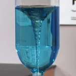 Pipeta com líquido azul vertendo em um copo de pé, ilustrando uma experiência científica ou uma demonstração de laboratório.