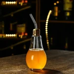 Lâmpada decorativa em forma de garrafa com bebida laranja, equipada com um canudo metálico, sobre uma superfície de madeira em um ambiente escuro e moderno.