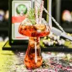 Copo de cocktail original em forma de balão de laboratório, decorado com ramos de pinheiro, perfeito para uma decoração de festa ou evento científico.