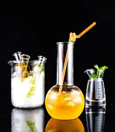 Vaso de laboratório contendo uma bebida laranja com um canudo, acompanhado de outros acessórios de laboratório em vidro, sobre fundo preto.
