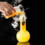Manipulação de uma experiência científica com um líquido laranja fumegante em um laboratório, usando um béquer e um tubo de ensaio, contra um fundo preto.