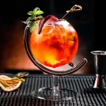 Cocktail colorido num copo original com guarnição de hortelã e fatia de limão, apresentado sobre um tapete preto com acessórios de bar ao fundo.