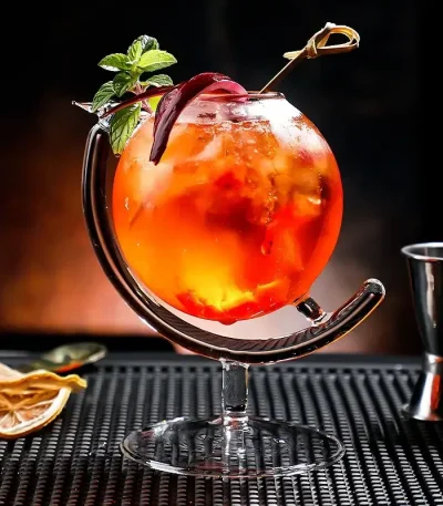 Cocktail colorido num copo original com guarnição de hortelã e fatia de limão, apresentado sobre um tapete preto com acessórios de bar ao fundo.