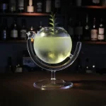 Cocktail luminoso num frasco científico com uma planta verde flutuante, apresentado sobre um suporte de vidro, ambiente de bar com prateleiras de garrafas ao fundo.