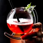 Cocktail colorido num copo de vidro transparente com guarnição verde, apresentado sobre um fundo preto.