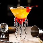 Copo de cocktail científico com decoração em forma de tentáculos em vidro transparente, preenchido com um cocktail amarelo-laranja com cereja, ambiente de bar moderno.