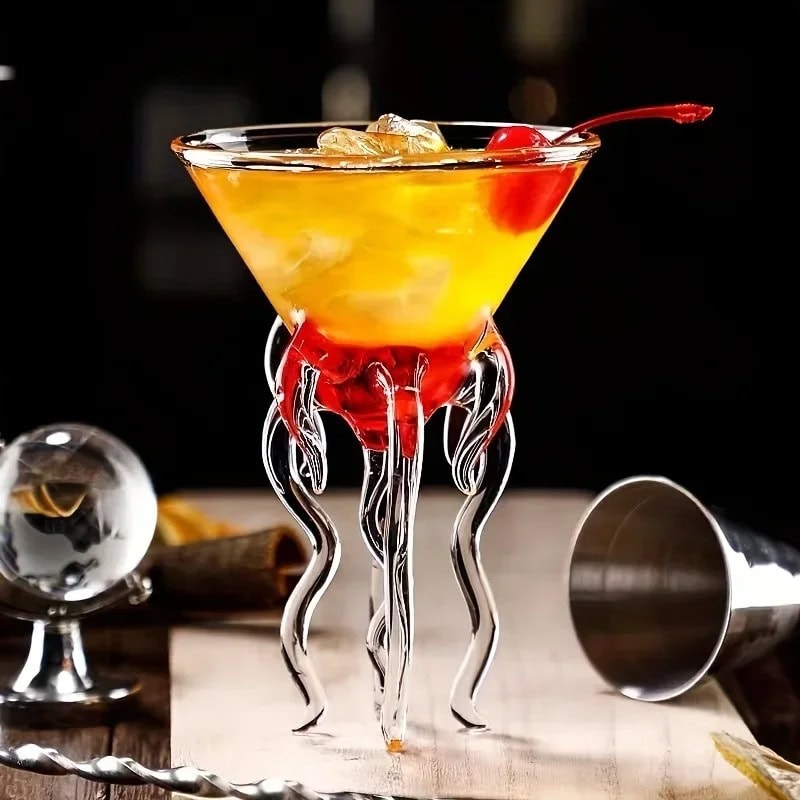 copo-de-cocktail-medusa-1 Copo de cocktail científico com decoração em forma de tentáculos em vidro transparente, preenchido com um cocktail amarelo-laranja com cereja, ambiente de bar moderno.