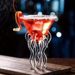 Cocktail vermelho em um copo em forma de medusa com tentáculos de vidro, guarnição de laranja e gelo, sobre uma superfície de madeira, ambiente noturno e misterioso.