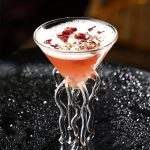 Cocktail rosa em um copo em forma de frasco com decorações florais.