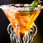 Copo de cocktail com guarnição de menta e limão, decorado com sal na borda, destacando uma bebida colorida e refrescante.