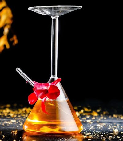 Béquer de laboratório com copo de cocktail suspenso, contendo um líquido laranja, decorado com uma flor vermelha, sobre uma superfície preta com brilhos dourados.
