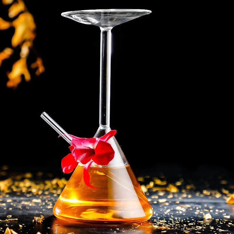 copo-de-cocktail-original-erlenmeyer-1 Béquer de laboratório com copo de cocktail suspenso, contendo um líquido laranja, decorado com uma flor vermelha, sobre uma superfície preta com brilhos dourados.