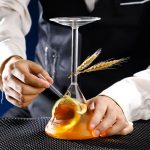 Frasco de laboratório com líquido laranja, decorado com trigo e uma colher, ilustrando a ciência e a pesquisa.