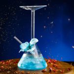 Vidros científicos contendo um líquido azul luminoso, com um balão de decantação e um tubo de vidro, sobre um fundo escuro com brilhos de luz e pétalas.