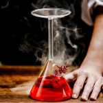 Frasco de laboratório de vidro contendo um líquido vermelho fumegante, colocado sobre uma superfície de madeira, com uma mão ao lado, ilustrando uma experiência científica.