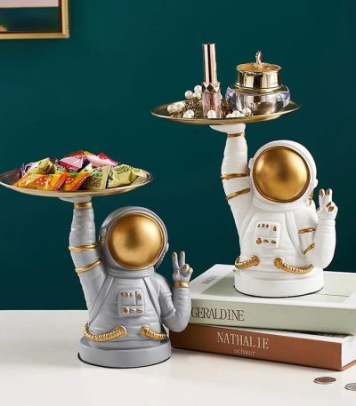 Figura de astronauta em cerâmica branca e dourada, usada como decoração, segurando uma bandeja com objetos de decoração e doces, estilo moderno e original.